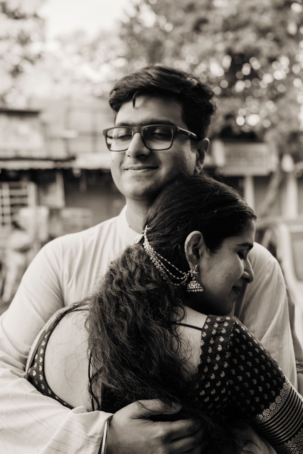 Black and white photo of a smiling young man with glasses embracing a woman in traditional Indian attire outdoors.
