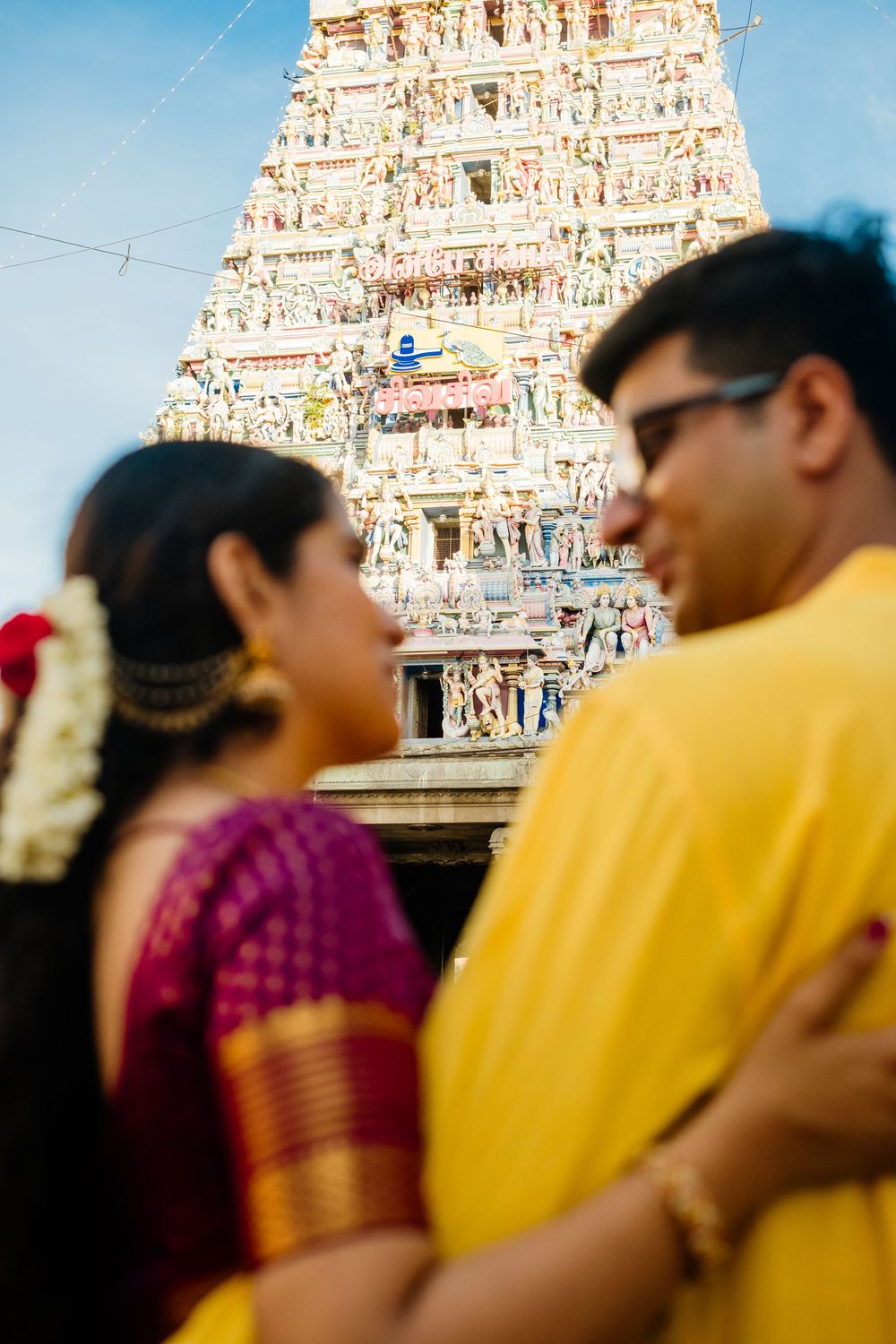 Indian couple in traditional attire embracing before ornate Hindu temple gopuram with colorful sculptures.
