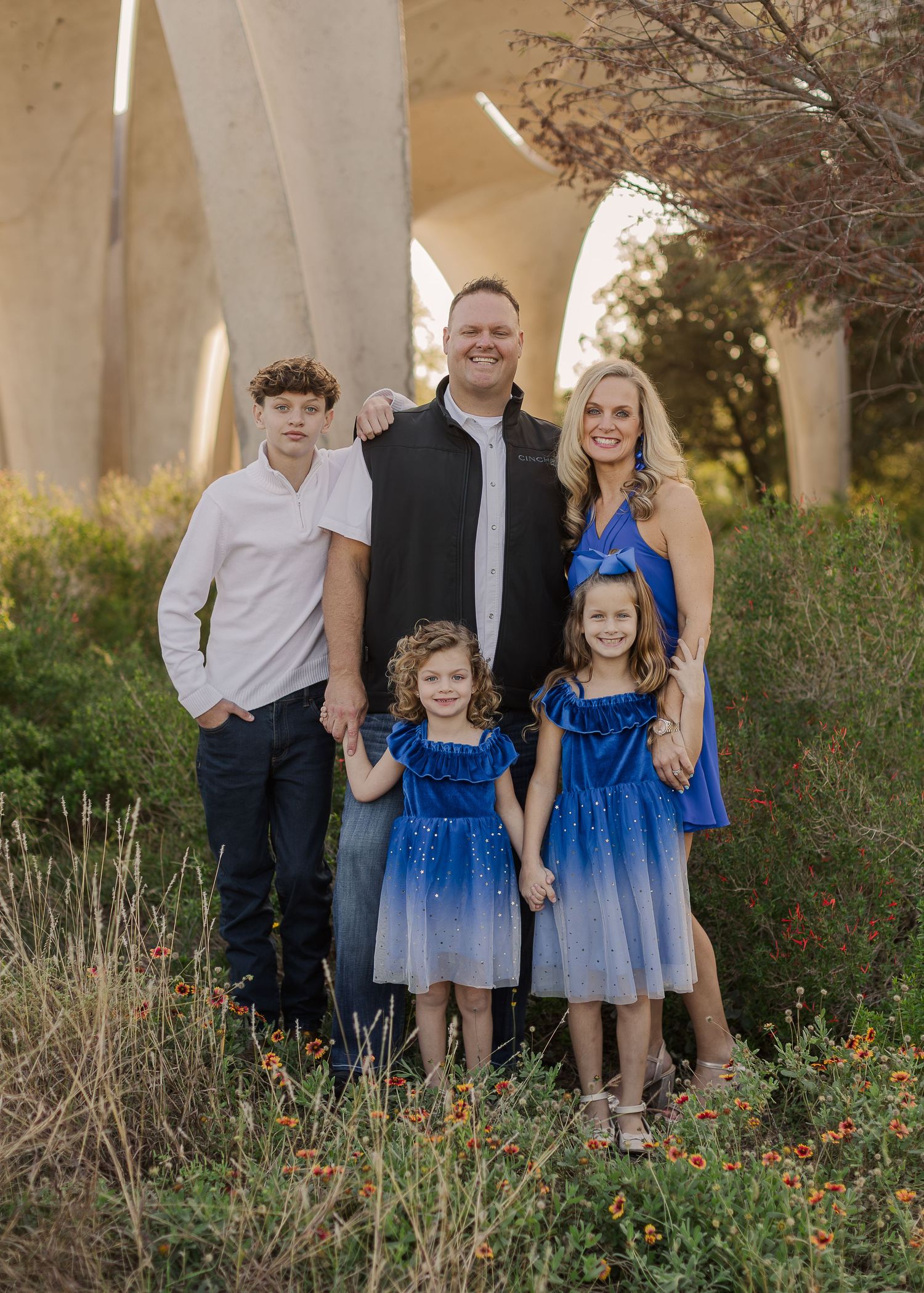 Family of five posing outdoors near concrete arches surrounded by wildflowers, wearing blue and white outfits.
