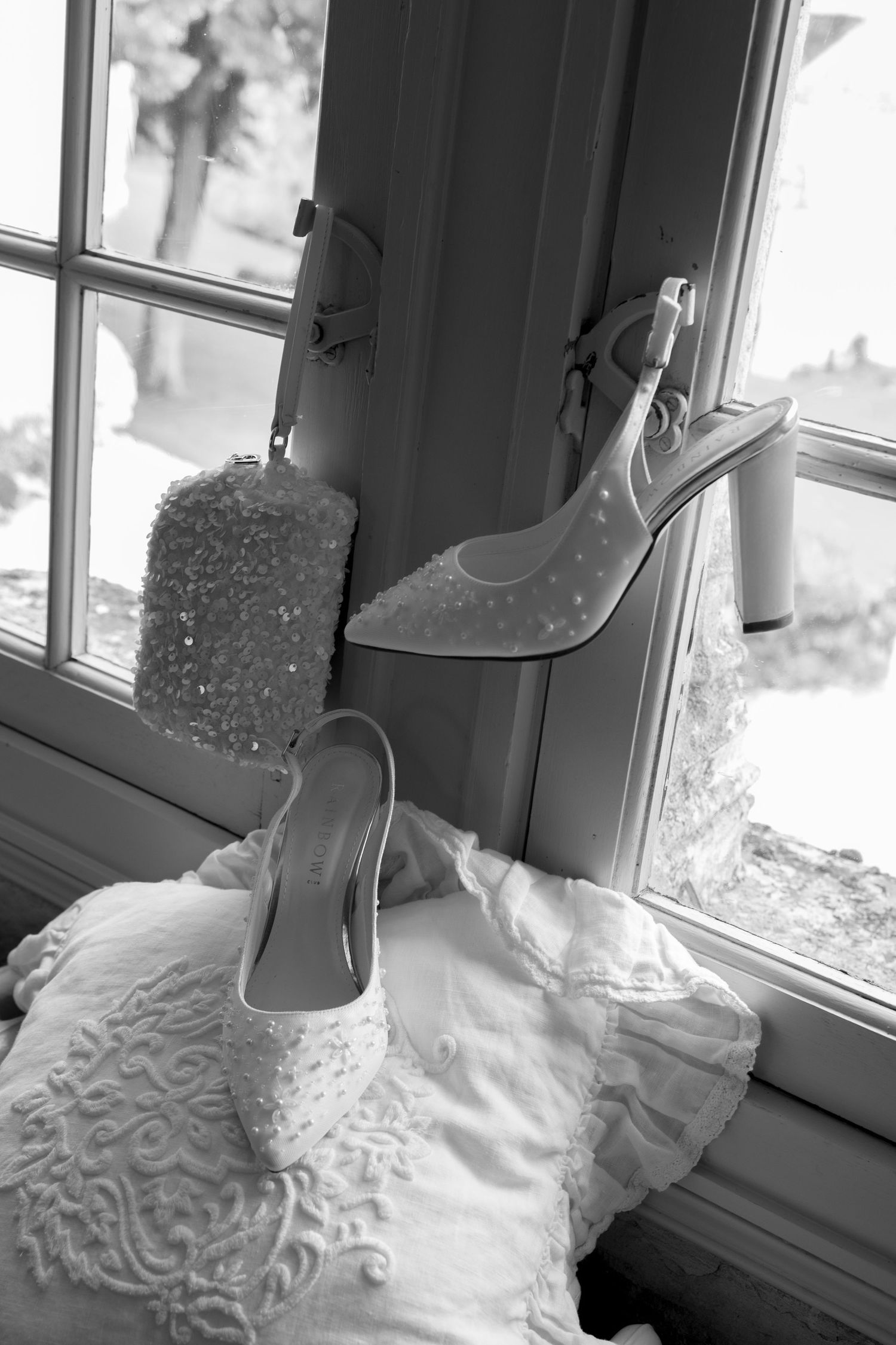 Black and white photo of elegant bridal heels and beaded clutch purse displayed on a lace wedding dress by a window.