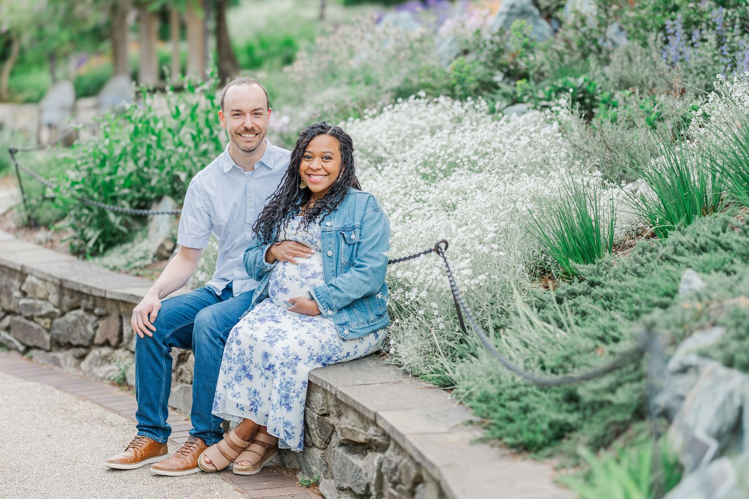 A couple sits together on a stone bench surrounded by lush garden flowers, the woman cradling a baby bump in a floral dress.