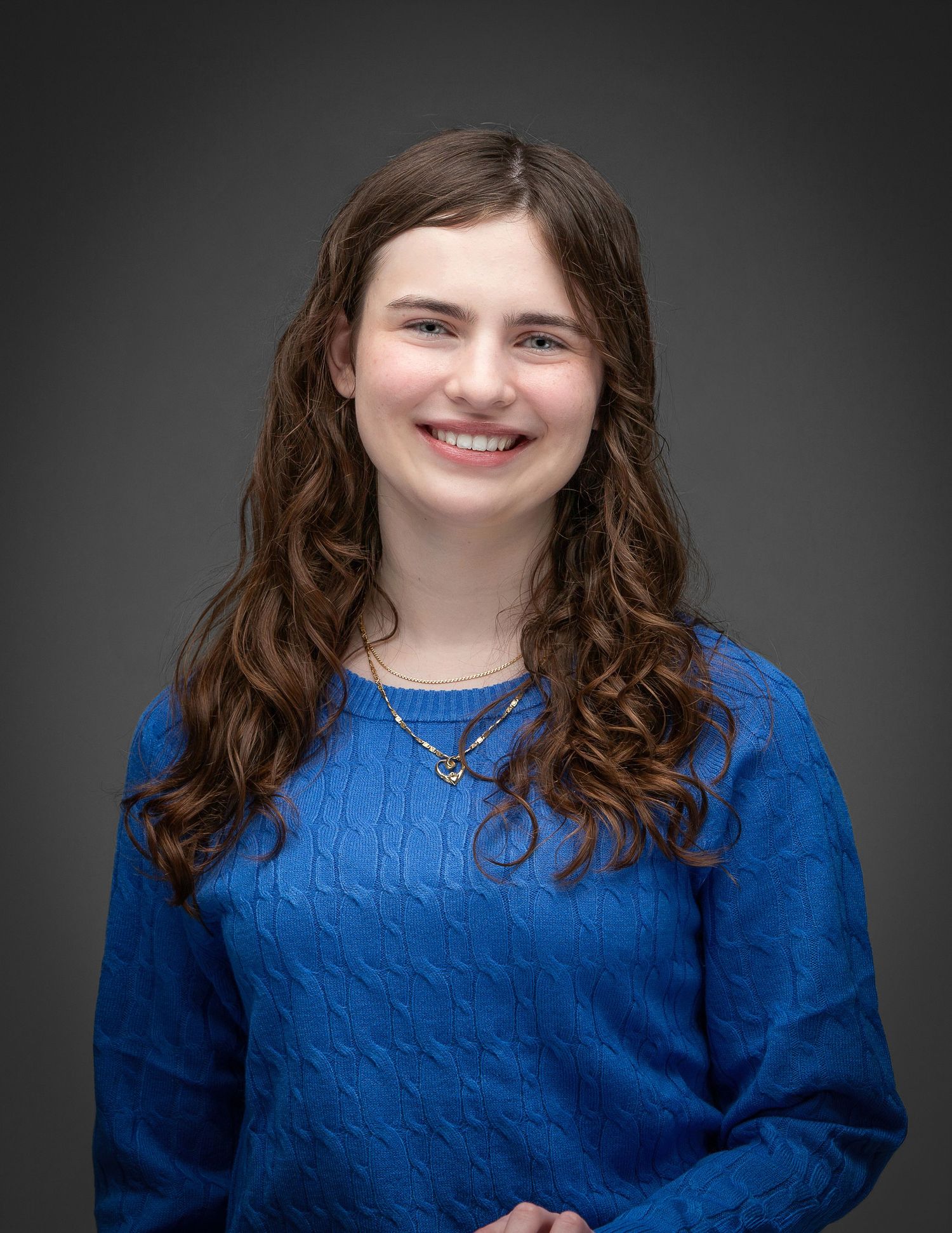 Young woman with curly brown hair smiling, wearing a blue cable-knit sweater and layered necklaces.