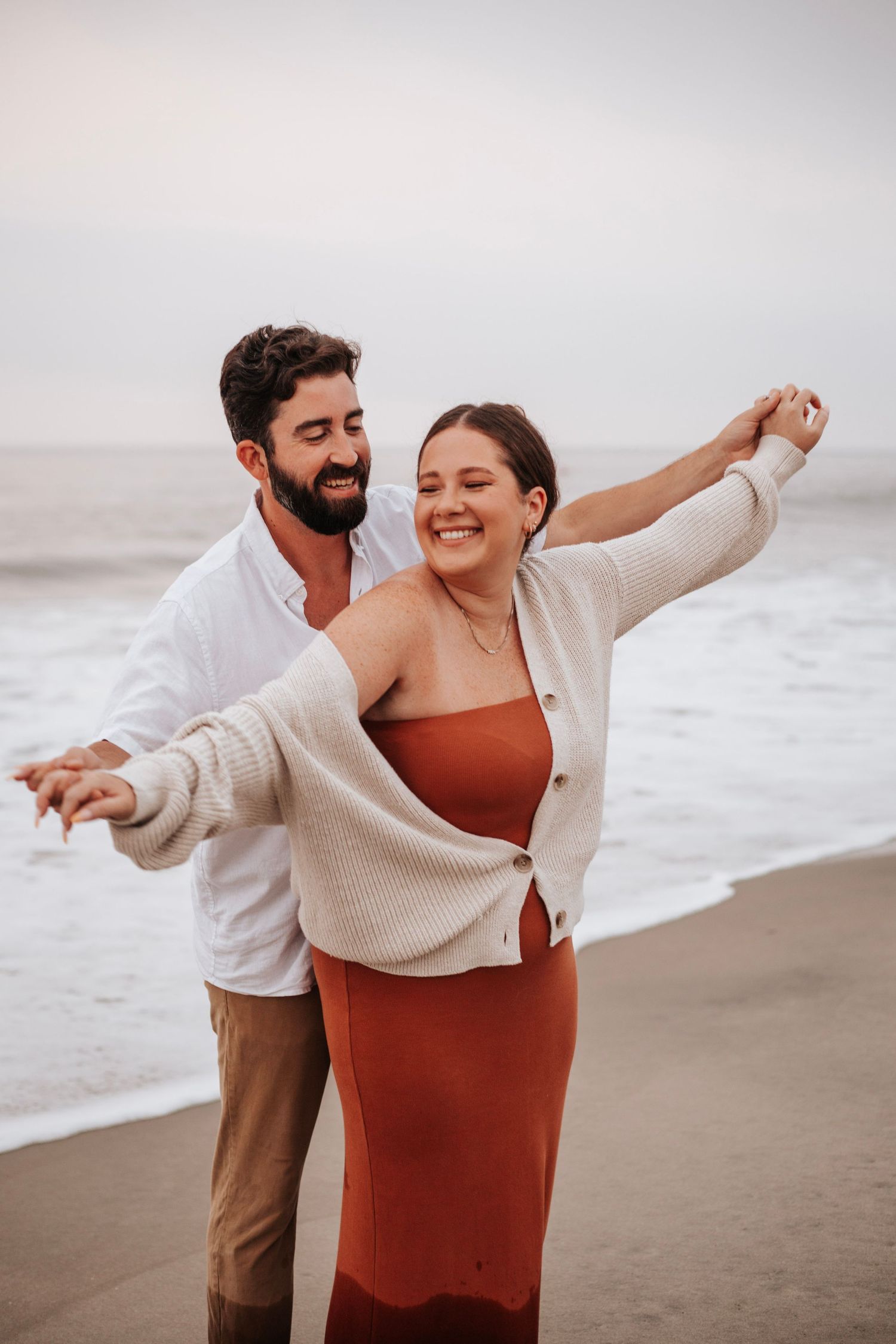 Happy couple dancing on a beach at sunset, woman in rust dress and man in white shirt smiling.