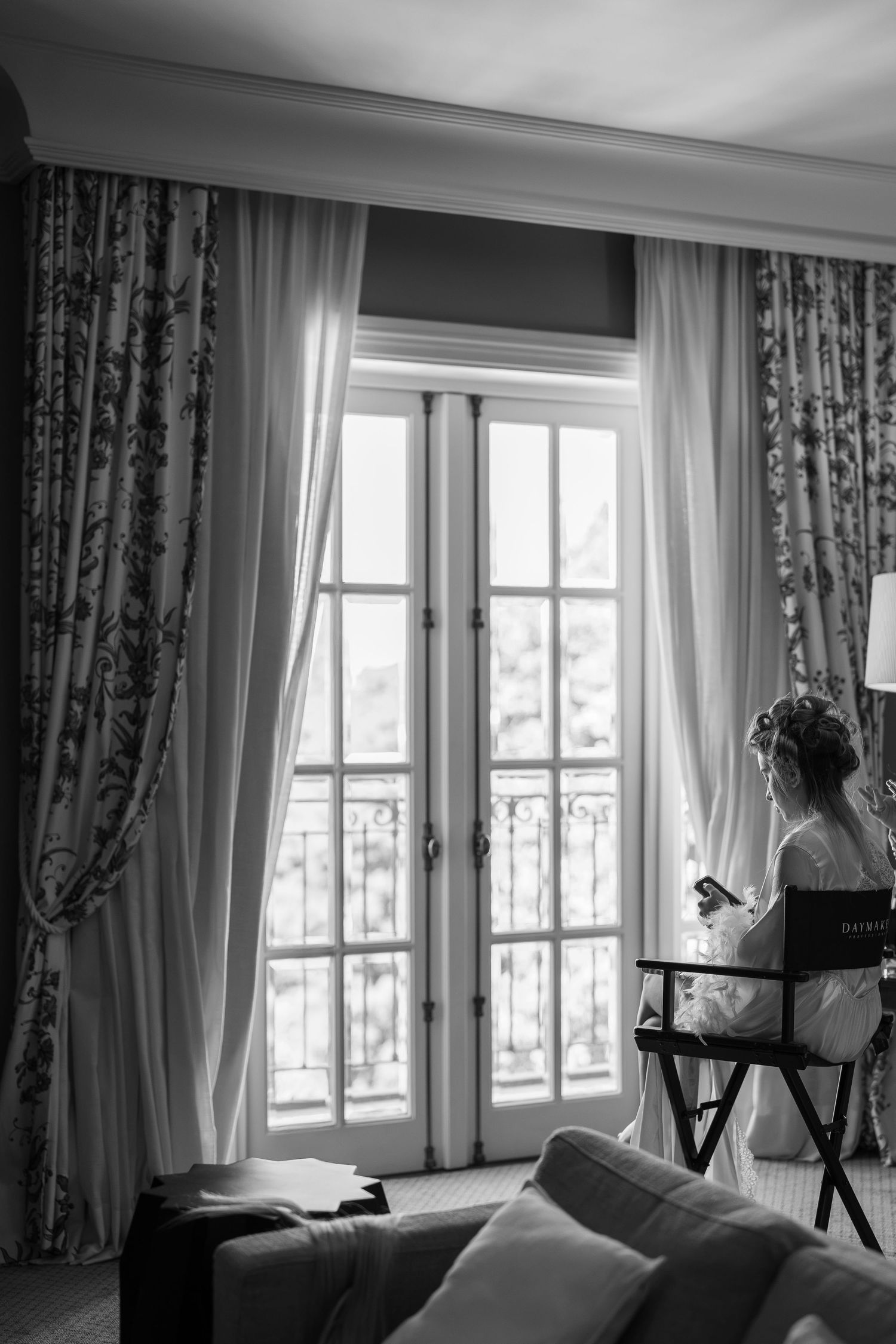Black and white photo of a bride sitting by elegant French doors with floral curtains during wedding preparations.