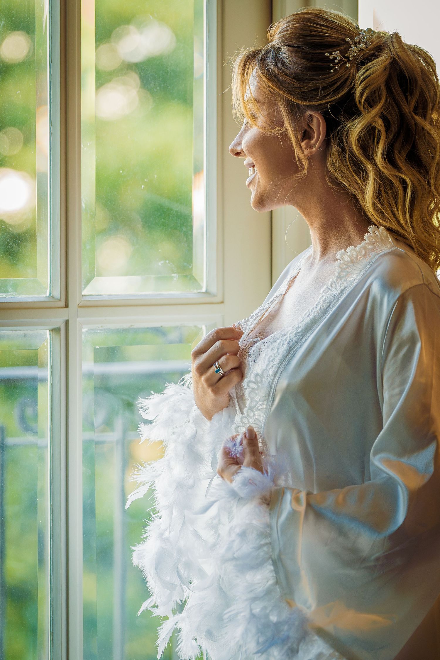 Smiling bride in white feather robe gazing out a sunlit window, wearing hair accessories and a ring.