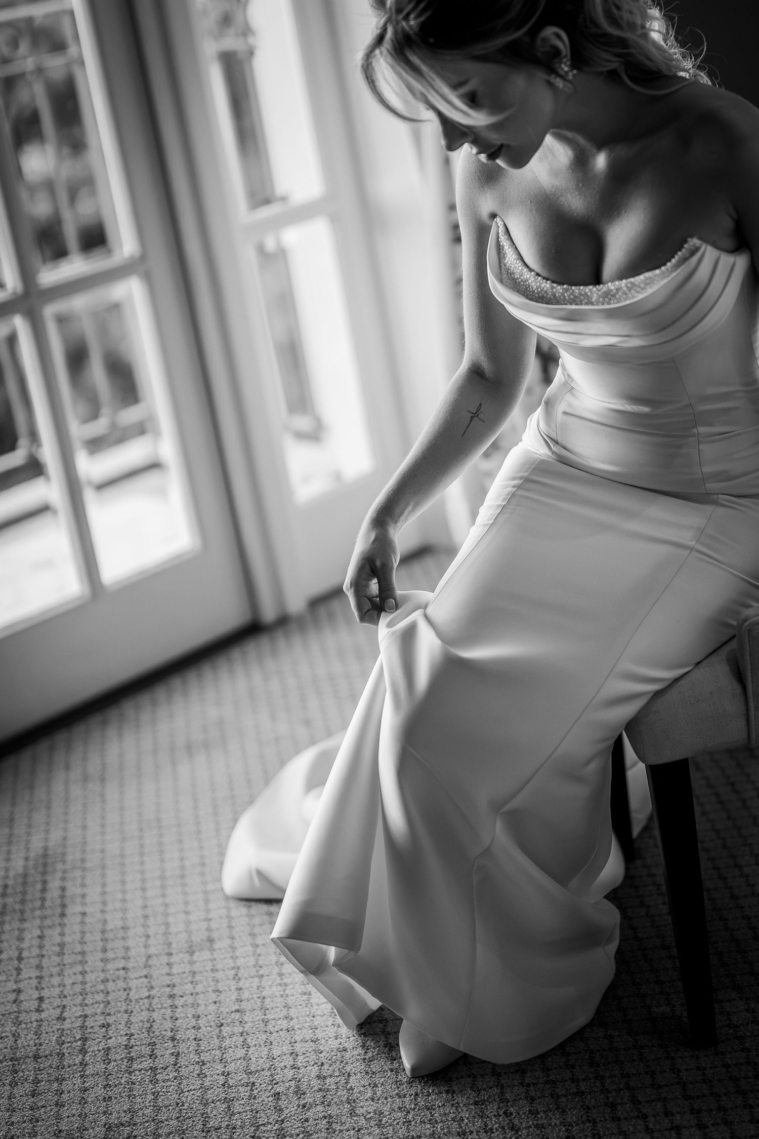 Black and white photo of a bride in an elegant strapless wedding gown, seated by a bright window, adjusting her dress.