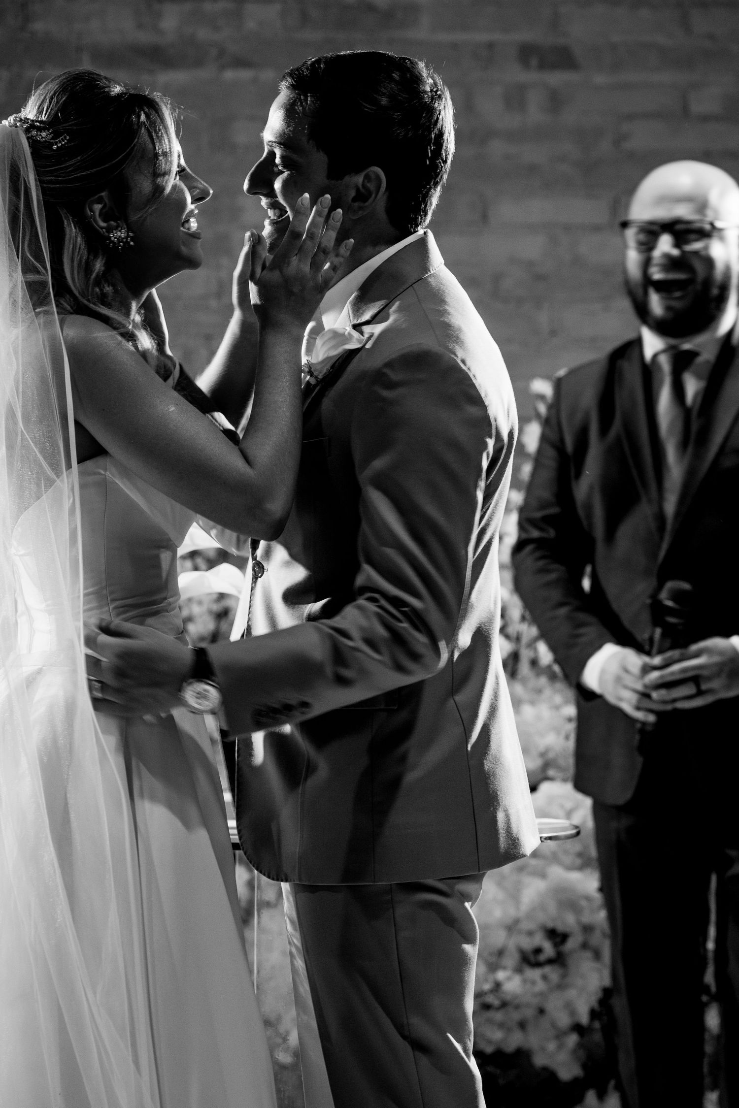 Black and white photo of bride touching grooms face at wedding ceremony as guest laughs joyfully in background.