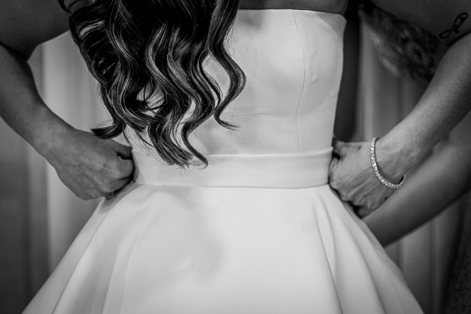 Black and white photo of a bride in a strapless wedding gown being helped with her dress by two assistants.