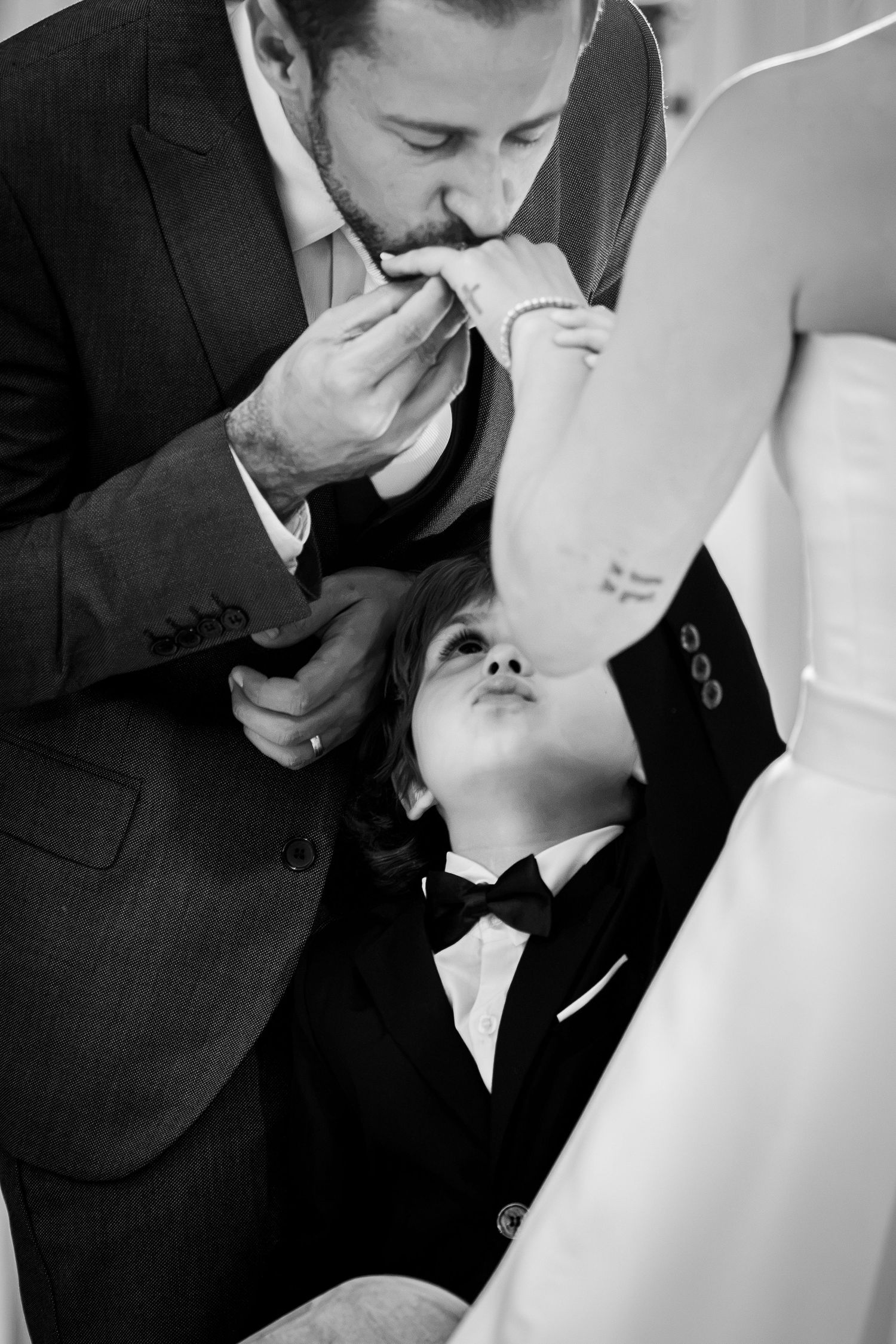 Groom in suit kisses bride's hand while young ring bearer in tuxedo looks up at them during wedding ceremony.