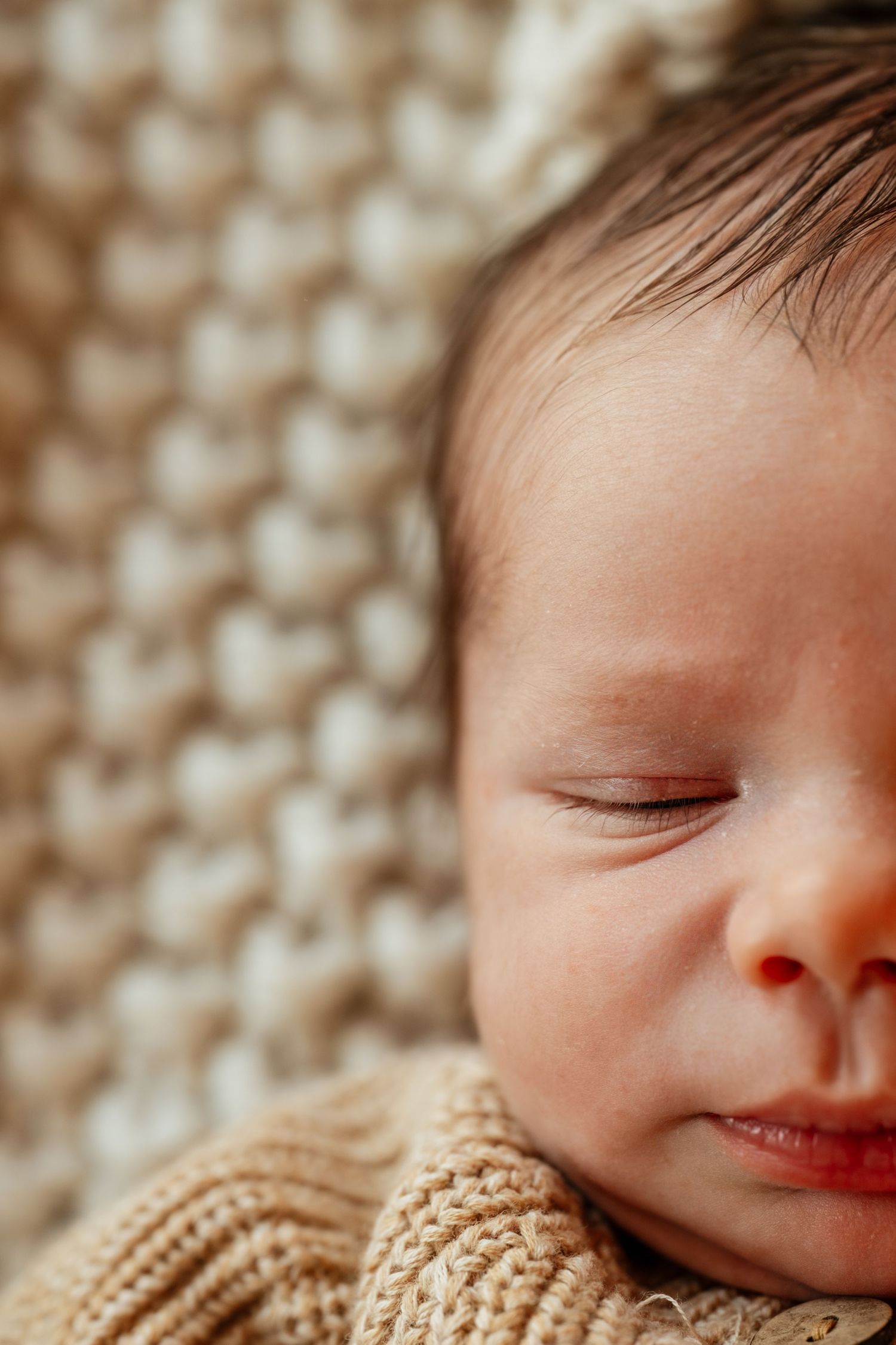 Close-up portrait of a sleeping newborn baby with dark hair, wearing a beige knit outfit on a cream textured blanket.