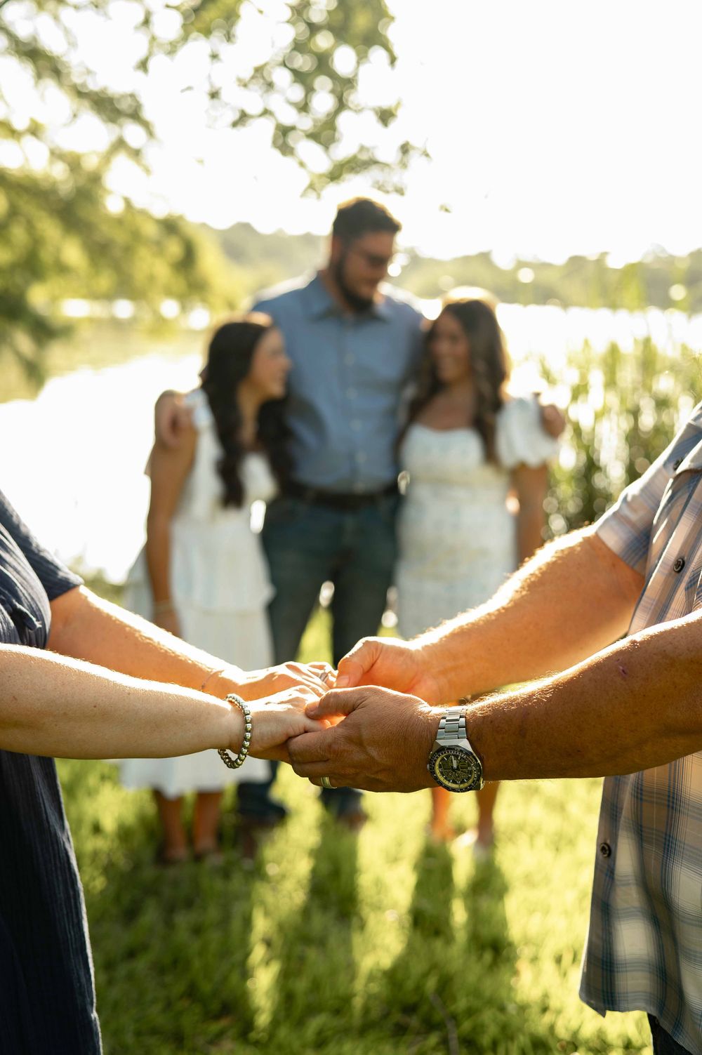 Two people hold hands in foreground while a couple stands near a sunlit lake surrounded by green trees.