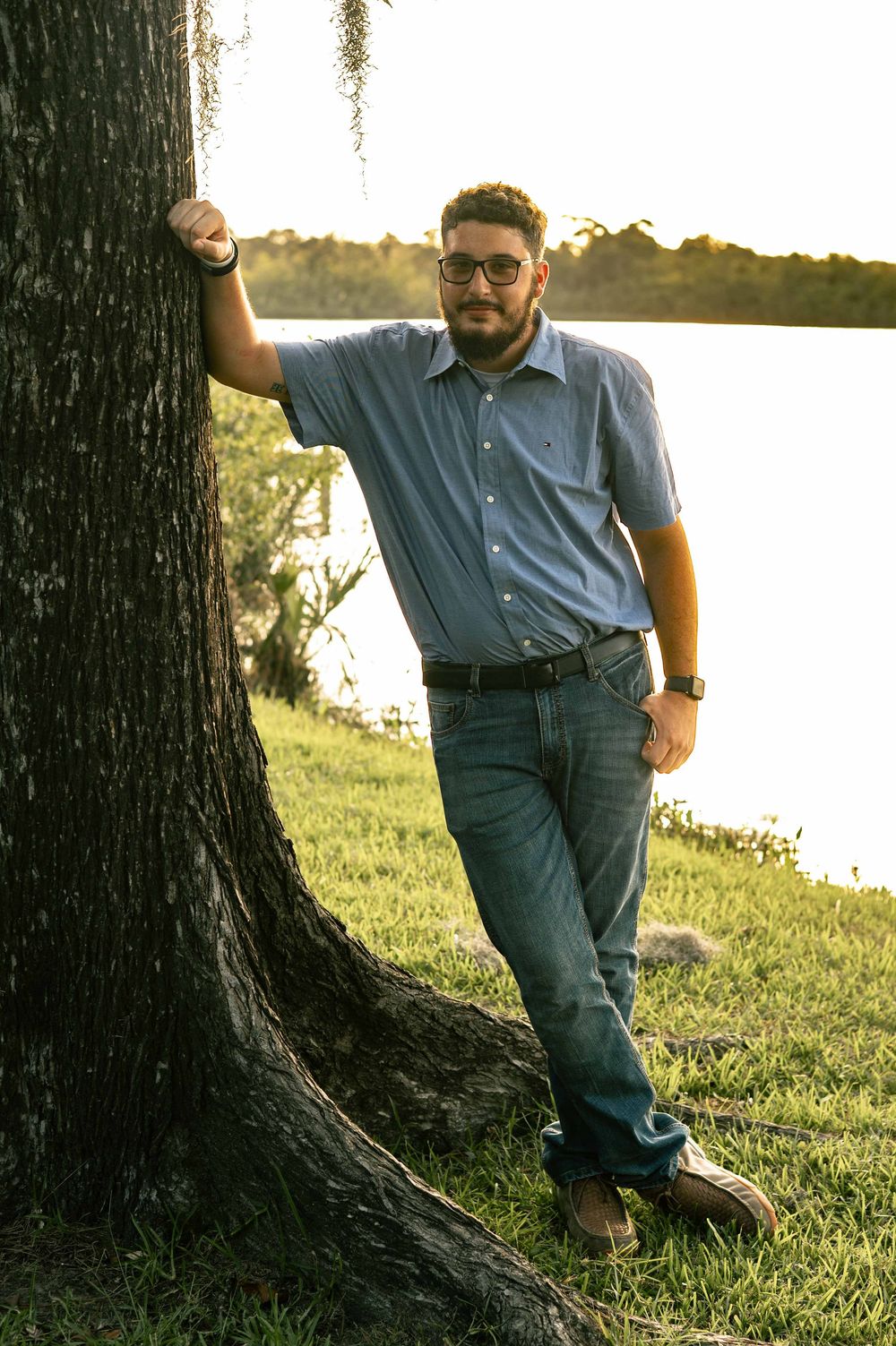 Man in blue shirt and jeans leaning against a tree by a lake at sunset, wearing glasses.