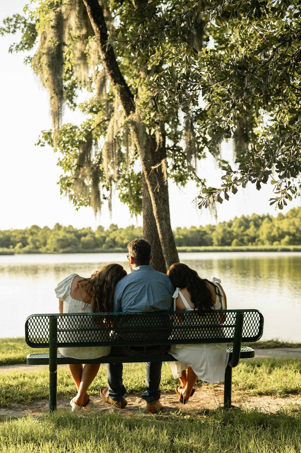 Three people sit together on a park bench under a moss-draped oak tree overlooking a calm lake.