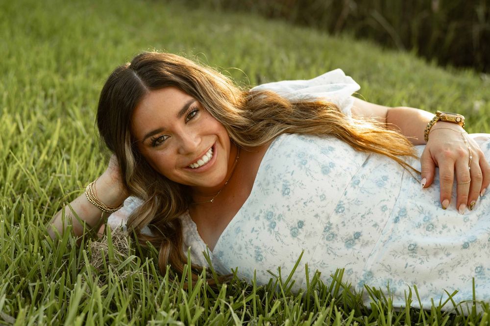 Smiling young woman in a floral dress lying on lush green grass, posing for a portrait photo outdoors.