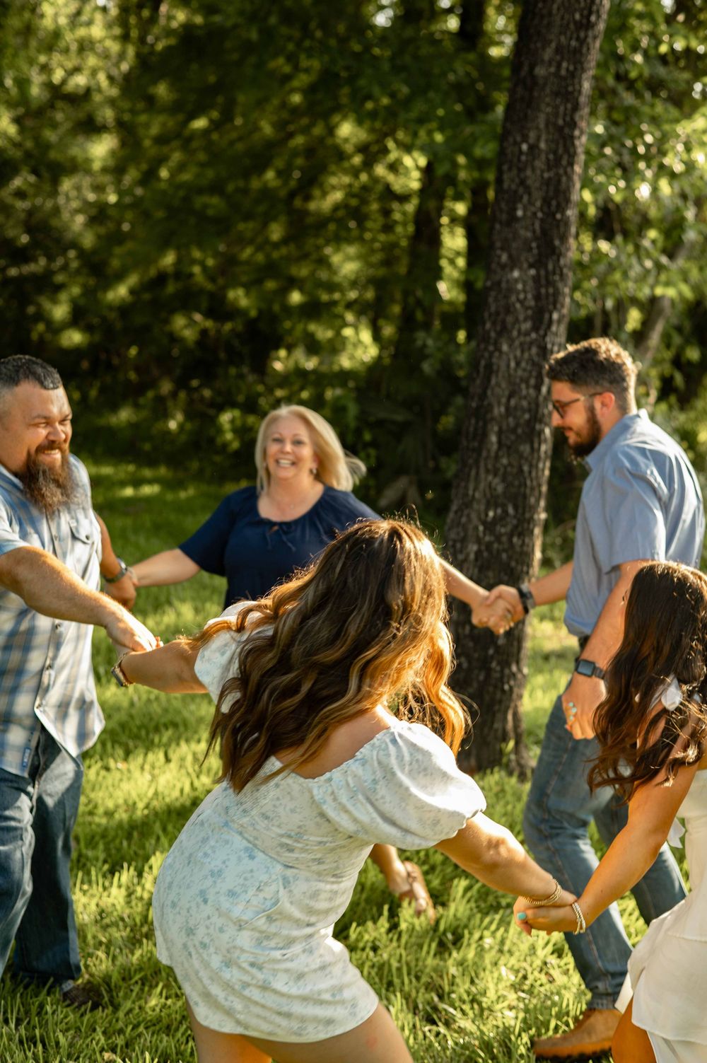 Family holding hands in a circle playing outdoors in a sunny green forest clearing.