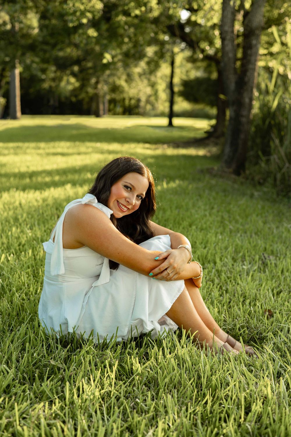 Young woman in white dress sitting on green grass in a sunny park, smiling warmly at the camera.