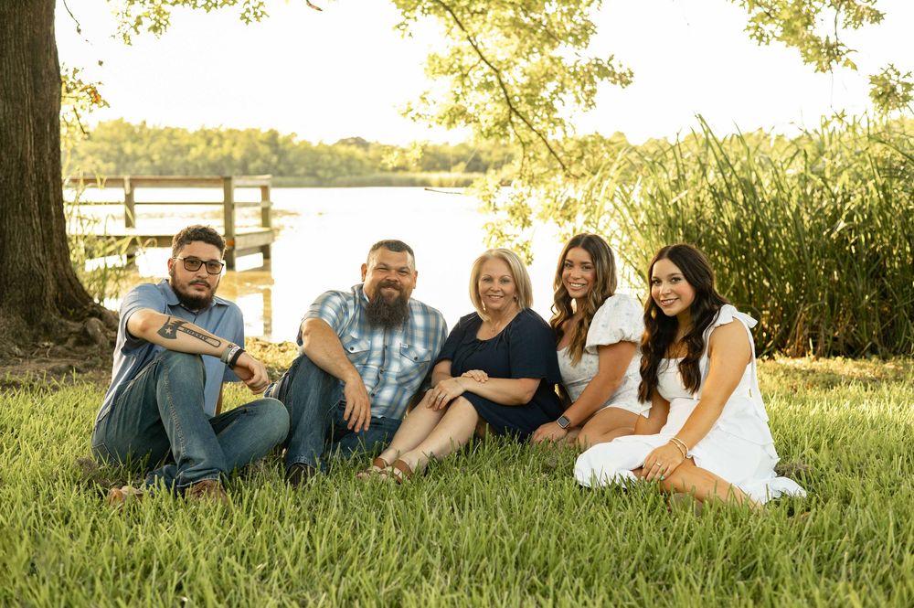Family of five smiling together on grass by a lake, posing for an outdoor portrait on a sunny day.