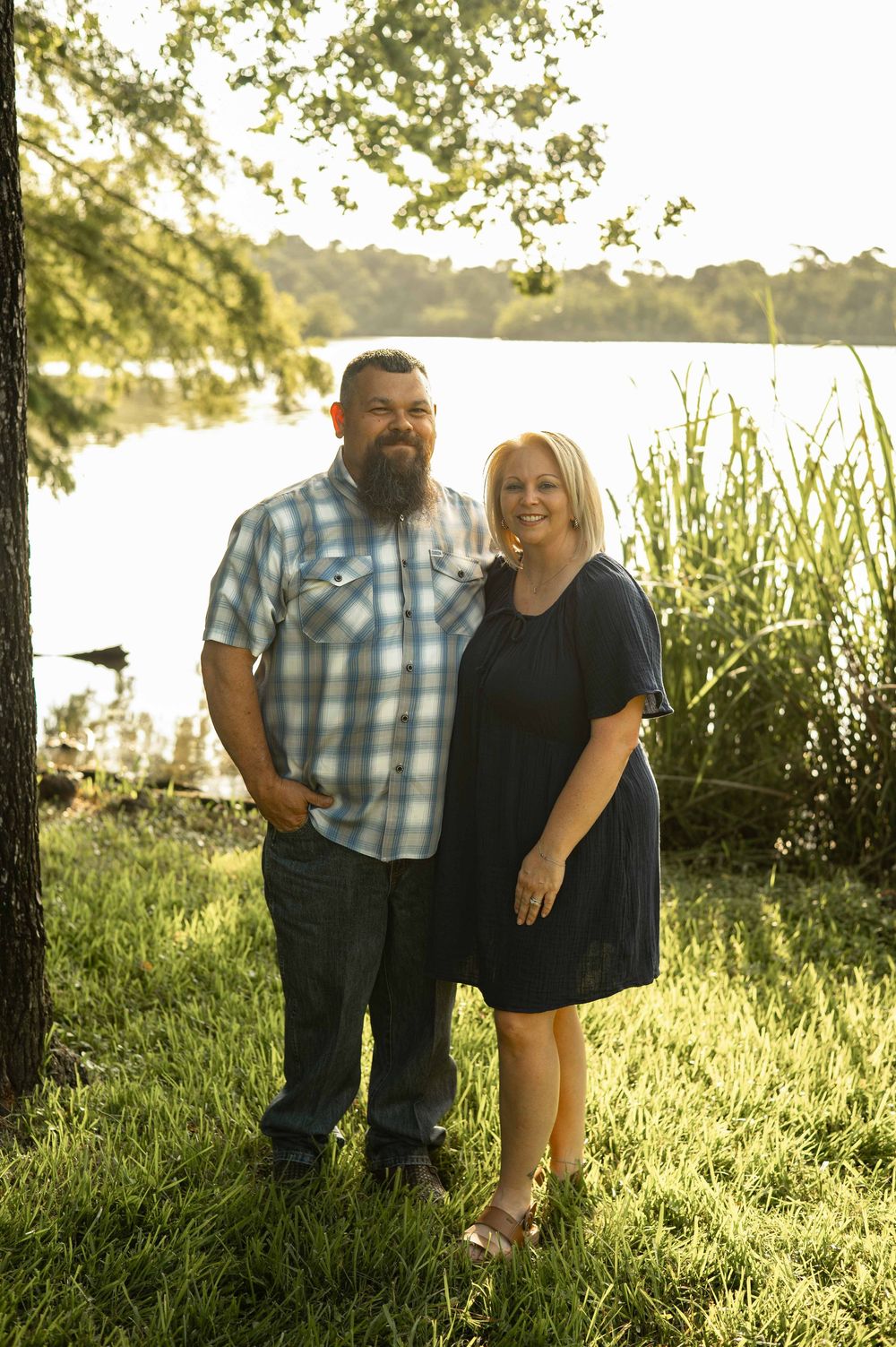 Couple smiling together outdoors near a sunlit lake, man in plaid shirt and woman in navy dress.