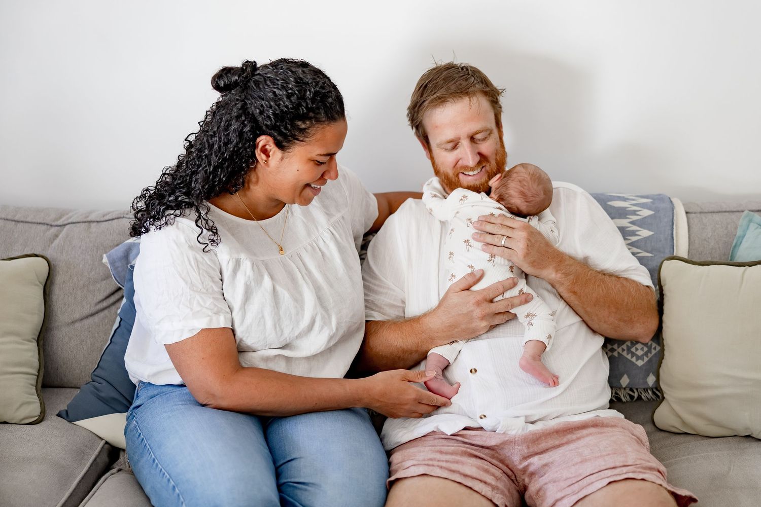 A happy couple sitting on a couch smiling at their newborn baby, with the father cradling the infant.