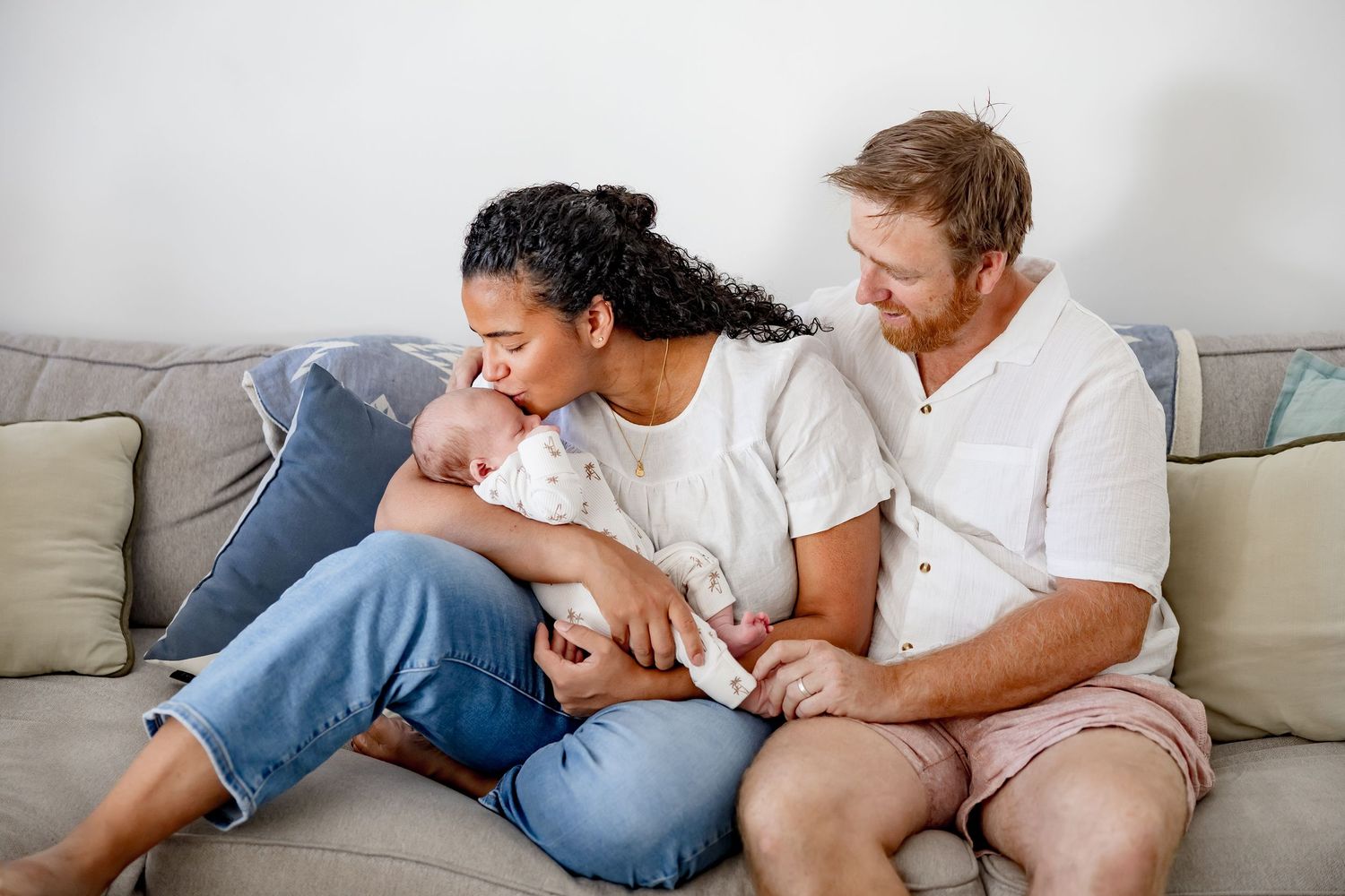 Happy interracial couple sitting on couch together admiring their newborn baby.