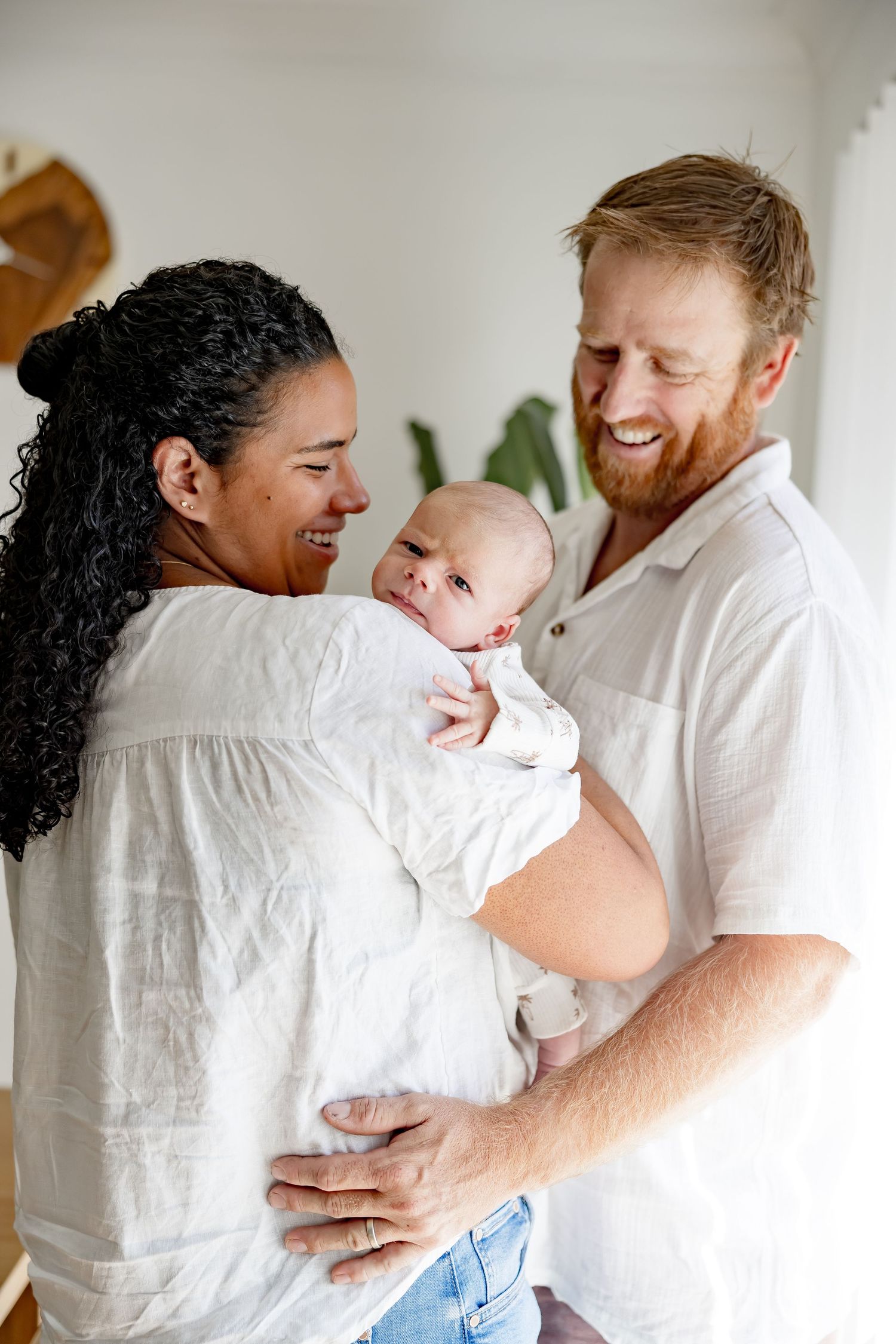 A smiling interracial couple stands together holding their newborn baby wrapped in a white blanket.