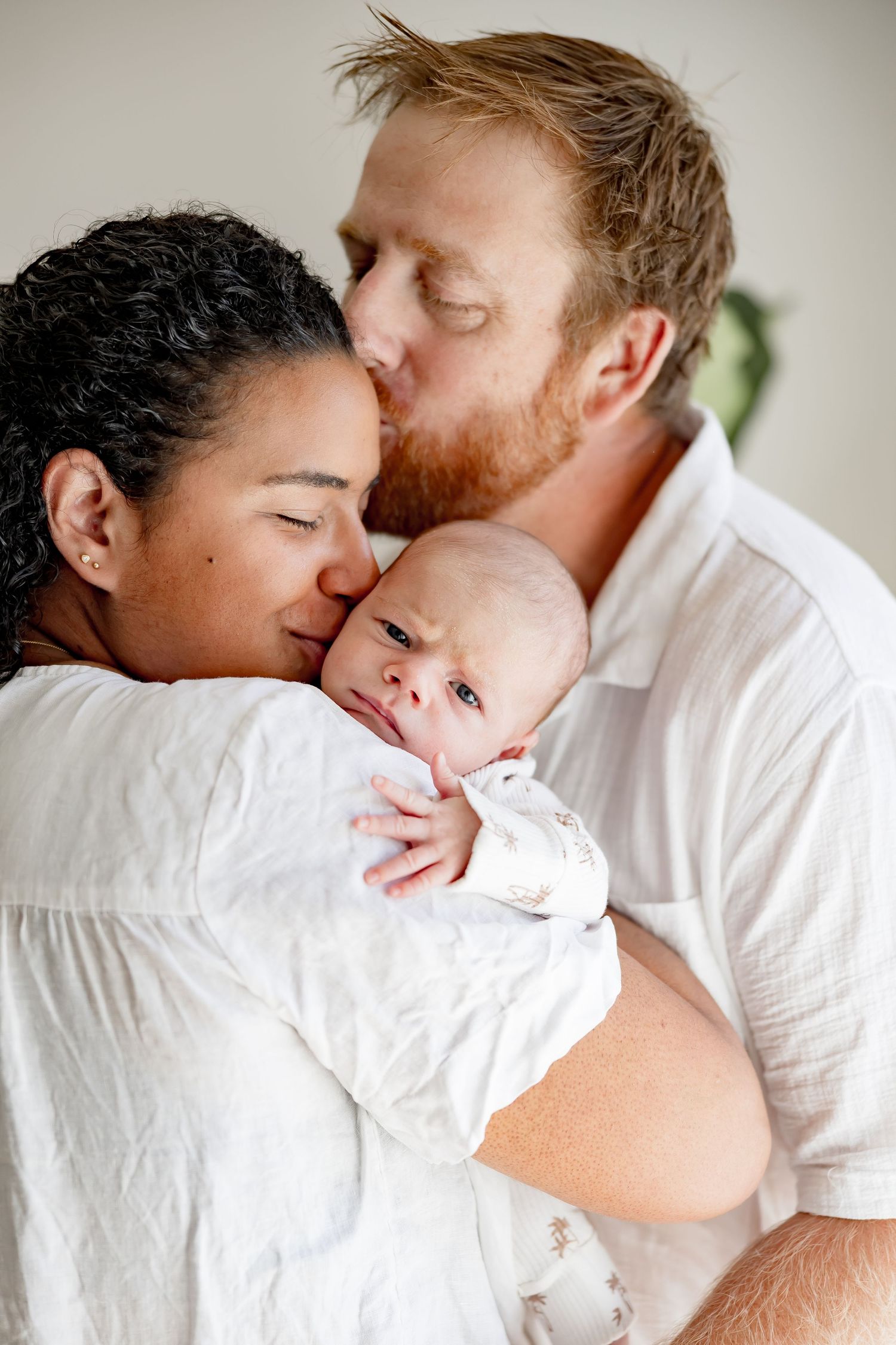 Interracial couple tenderly kissing and cuddling their newborn baby dressed in white.