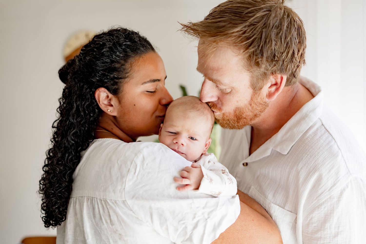 Interracial couple kissing their newborn baby dressed in white.
