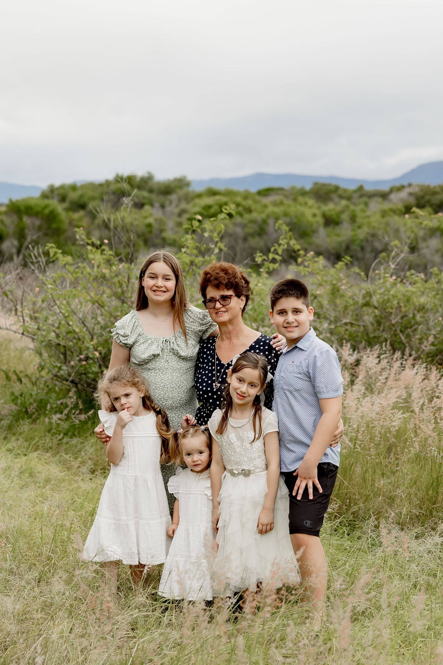 Grandmother posing with four grandchildren in a grassy field with green shrubs and mountains behind.
