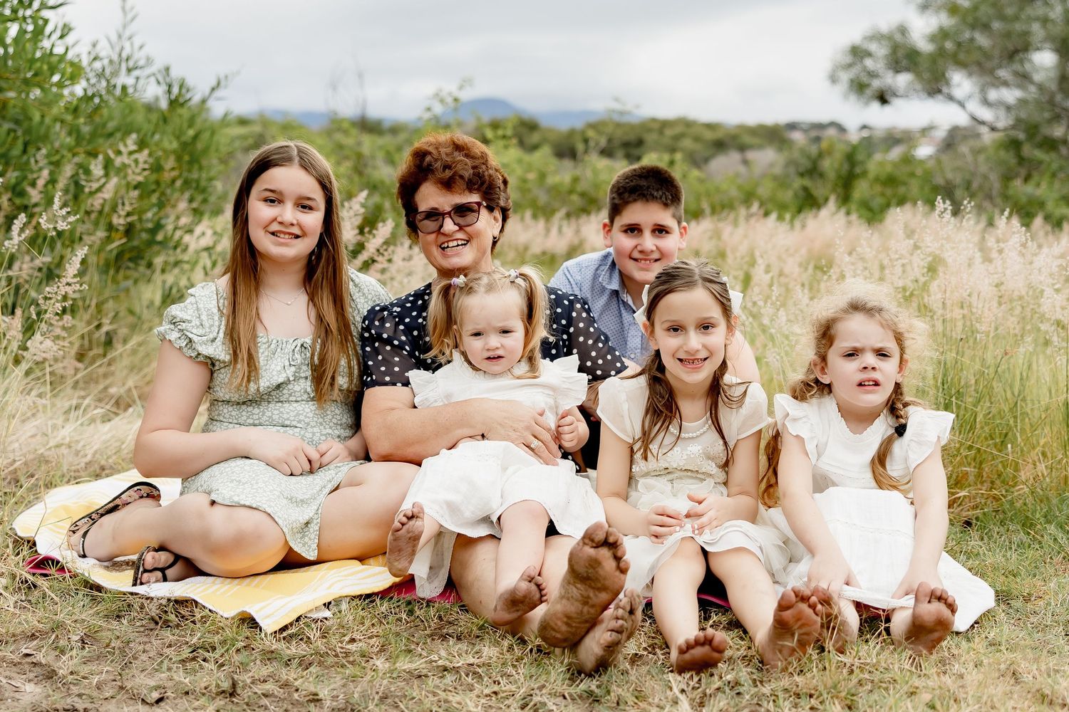 A grandmother sits outdoors in a grassy field with five young grandchildren, smiling together for a family portrait.