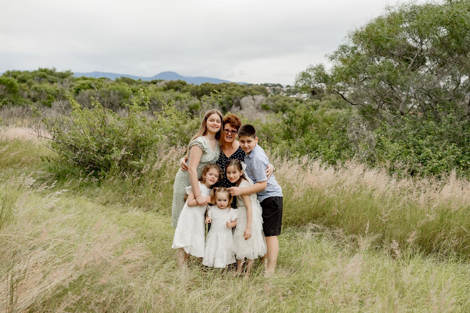 Family of six posing together in a lush green meadow with mountains in the background.