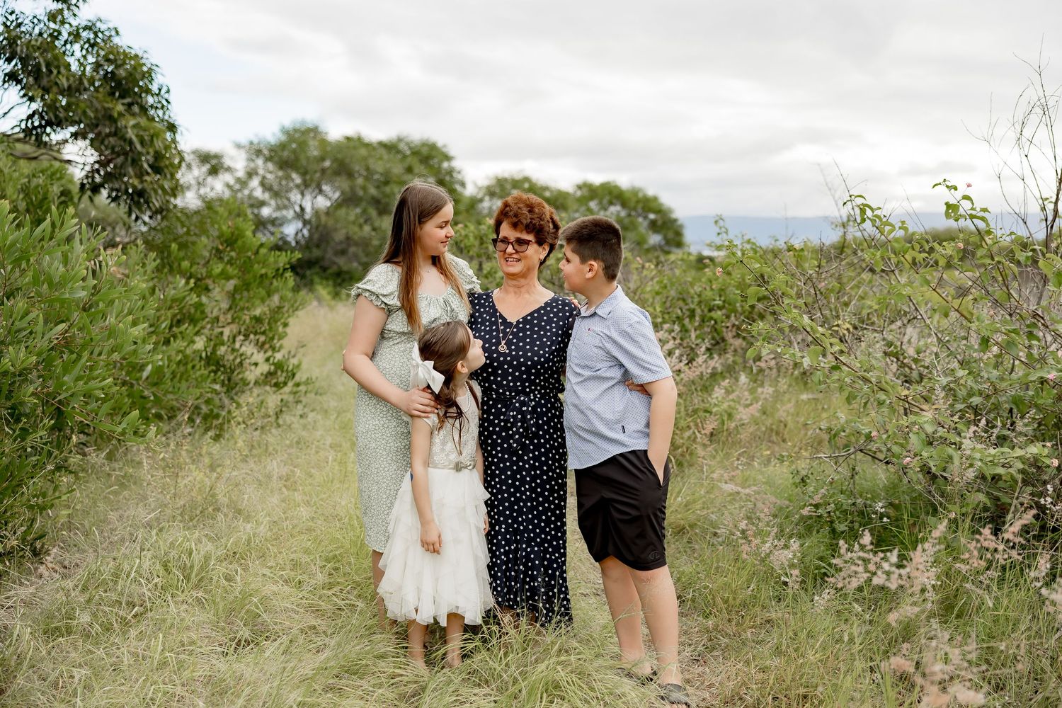 Family of three posing in a lush green meadow path, mother with two children smiling outdoors on a cloudy day.