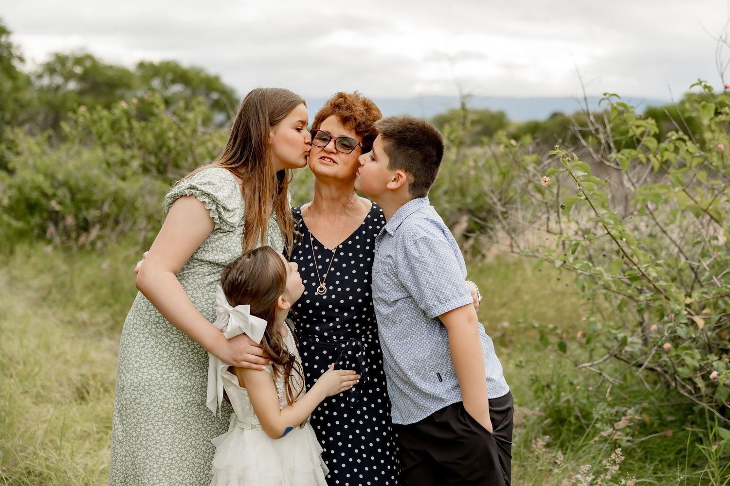 Two teenagers kissing their grandmother on the cheeks outdoors in a green natural setting.