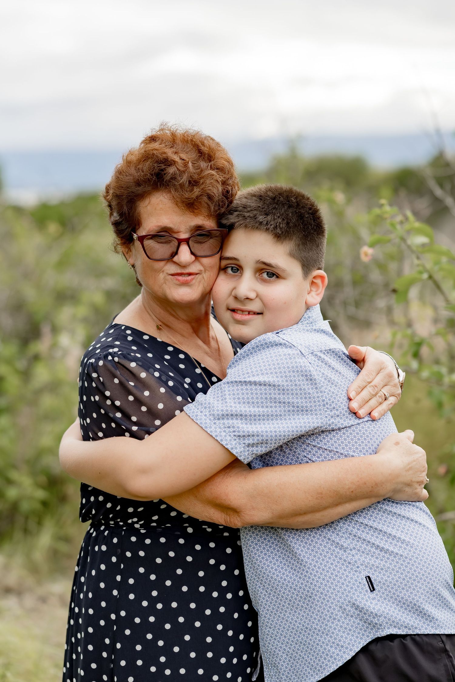 Grandmother with red curly hair hugging a young boy outdoors with green foliage in background.
