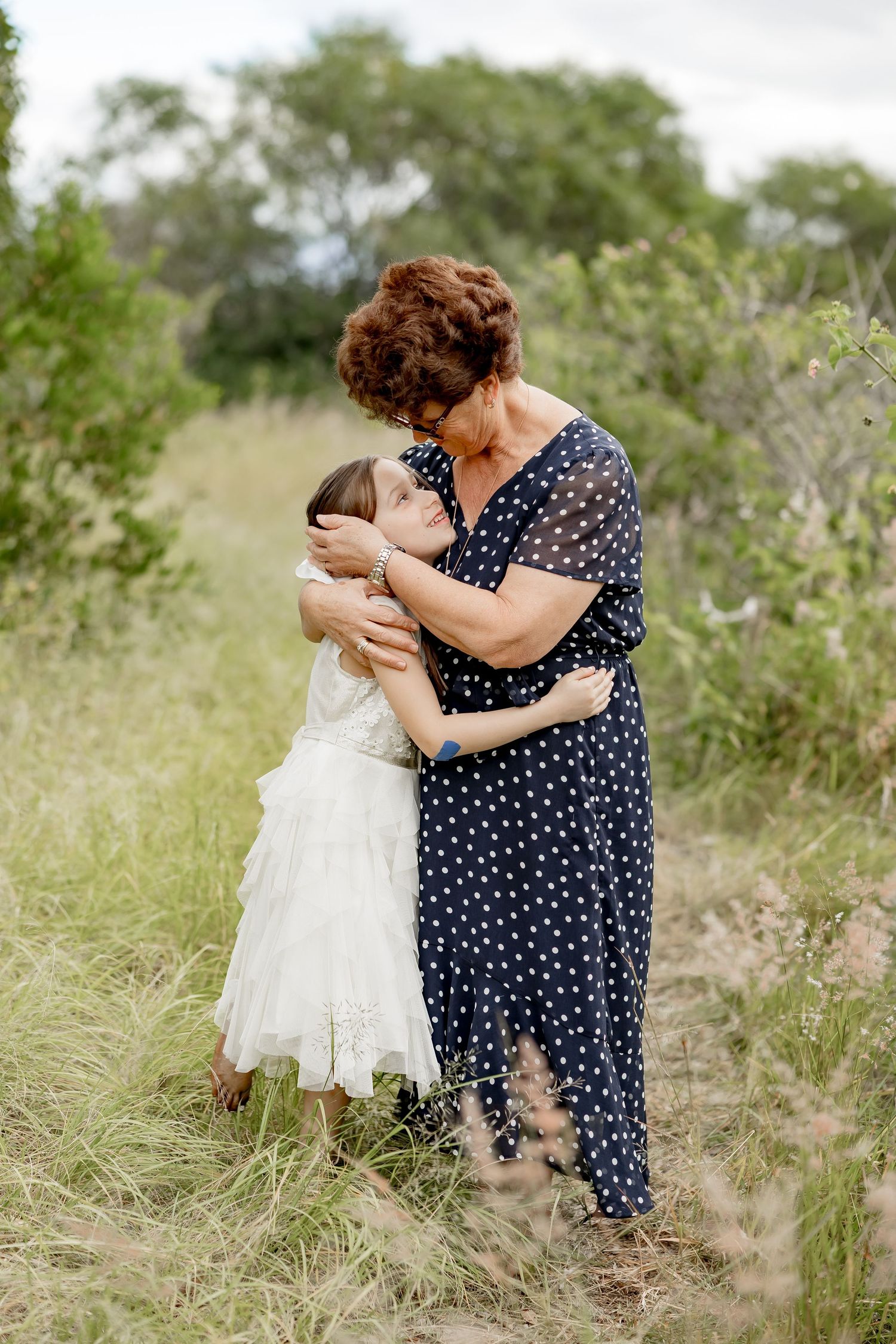 A grandmother tenderly embraces her granddaughter on a nature path surrounded by tall grass and green trees.
