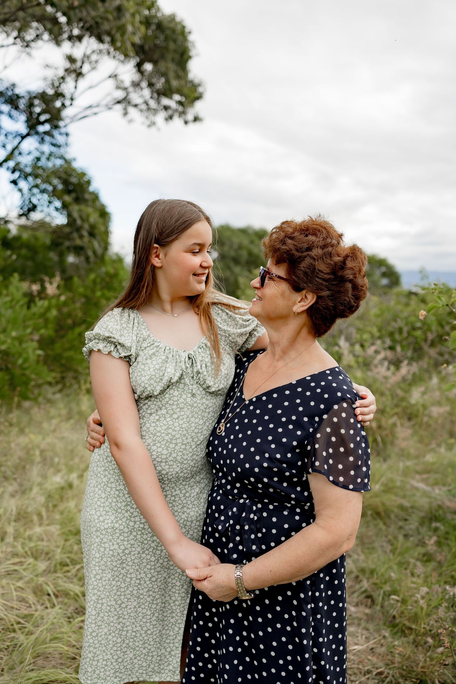 A teenage girl and older woman in floral dresses embrace warmly outdoors in a grassy field.