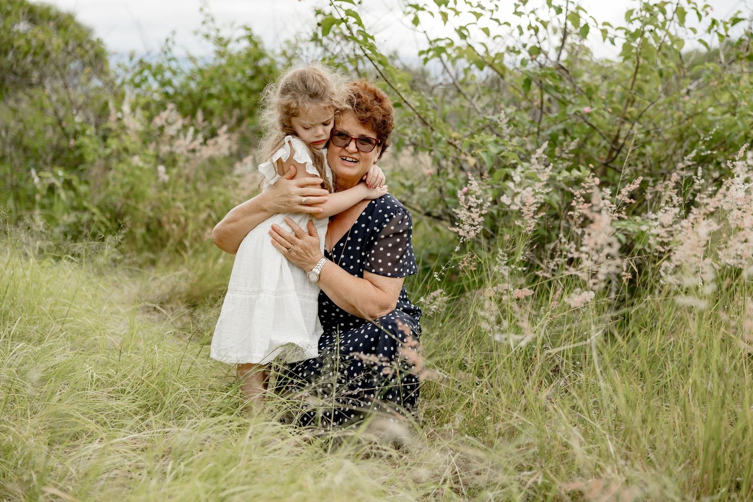 Grandmother hugging young granddaughter in white dress outdoors in lush green field with wildflowers.