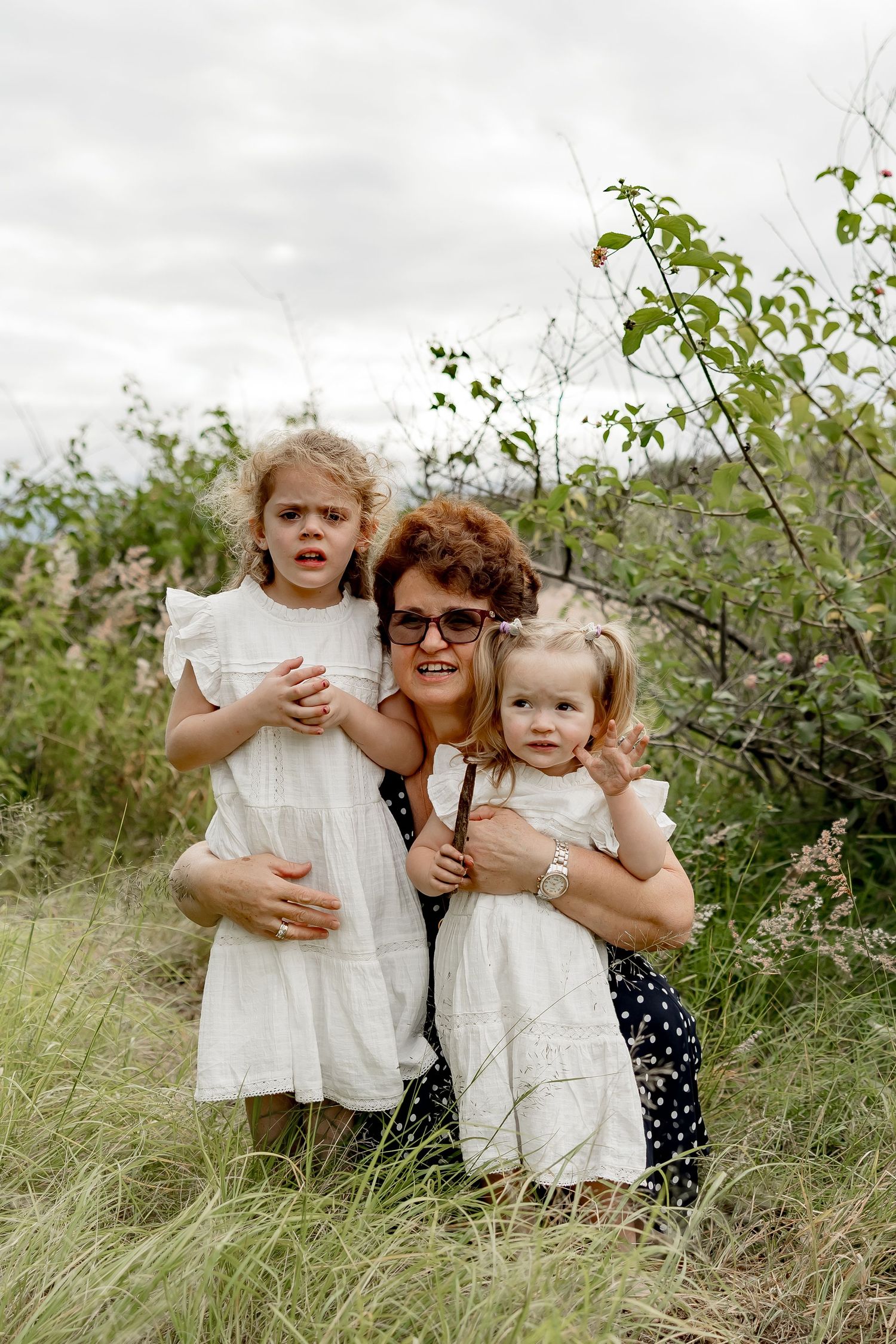 Grandmother with curly red hair and glasses hugs two young girls in white dresses outdoors in tall grass.