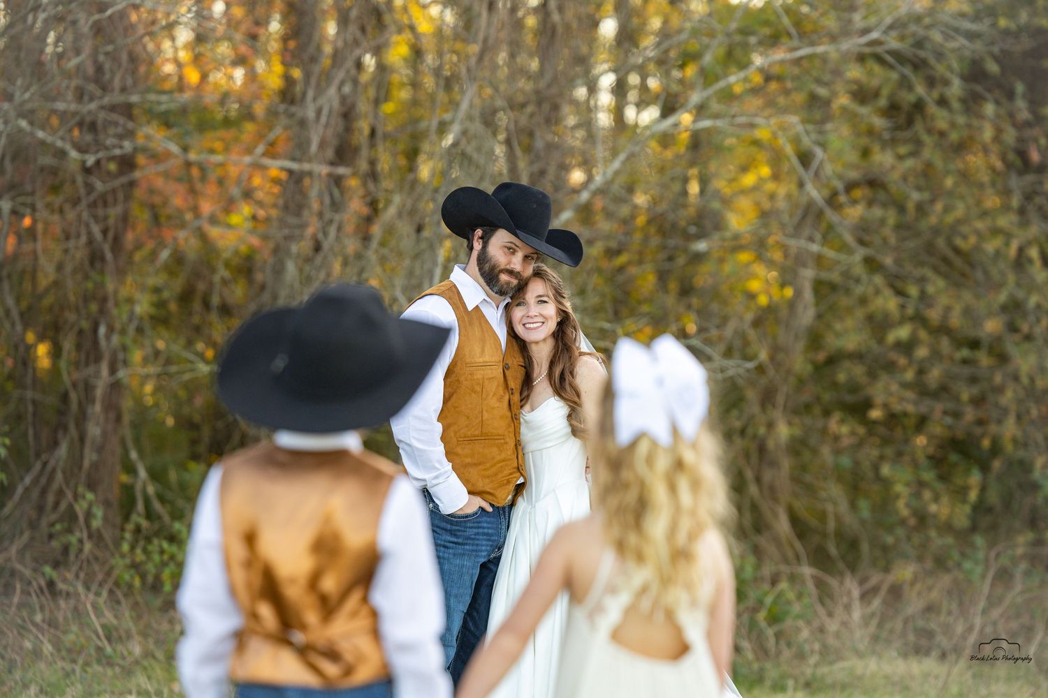 Rustic wedding couple in cowboy hats pose outdoors among autumn foliage, with guests in foreground.