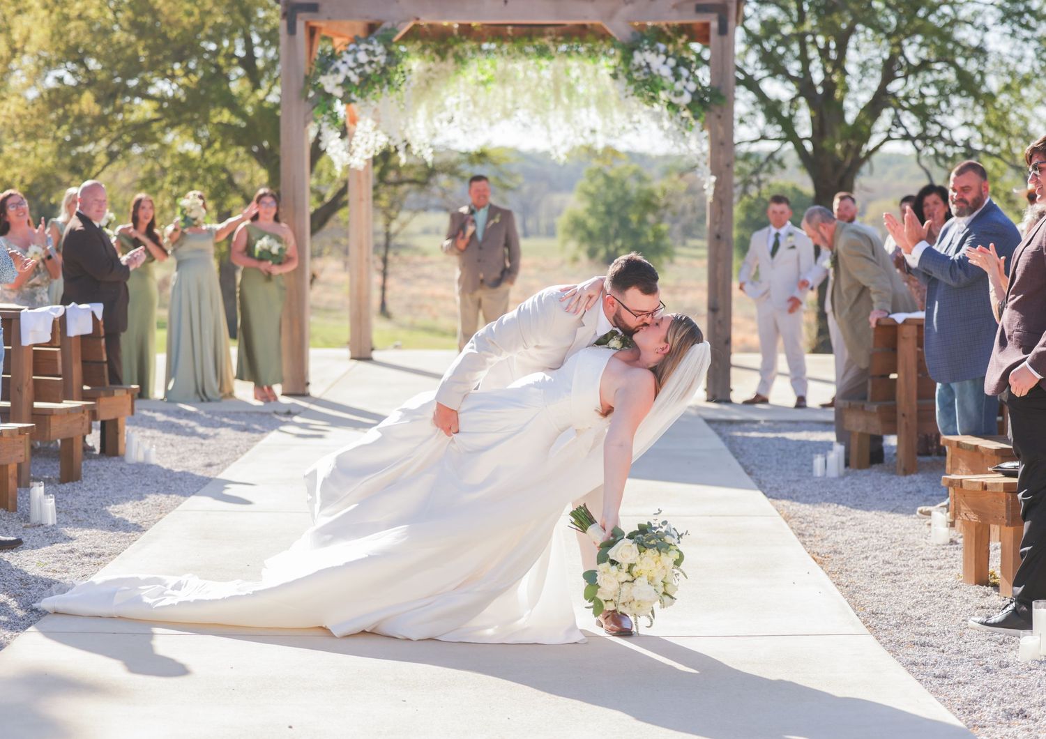 Bride and groom share a romantic dip kiss at outdoor wedding ceremony with guests applauding along aisle.