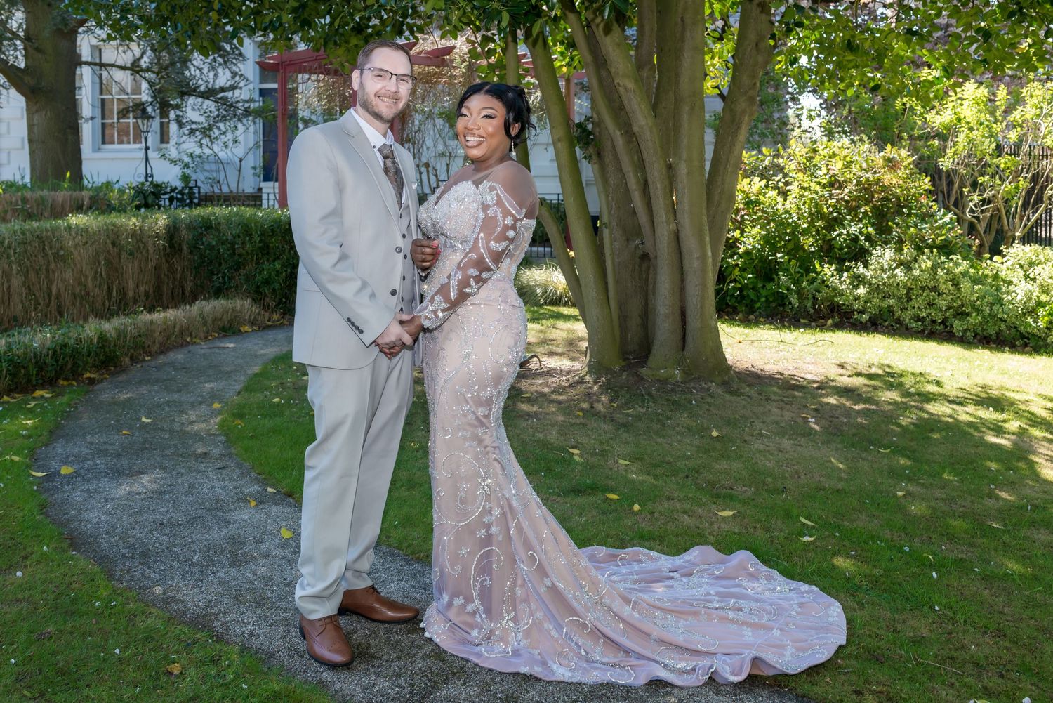 Bride in lace gown and groom in gray suit pose together in a garden on their wedding day.
