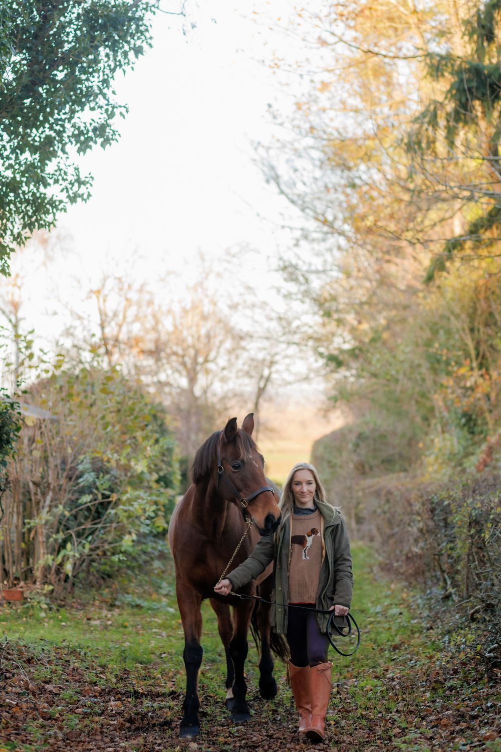 A young woman leads a brown horse along an autumn woodland path surrounded by colorful foliage.