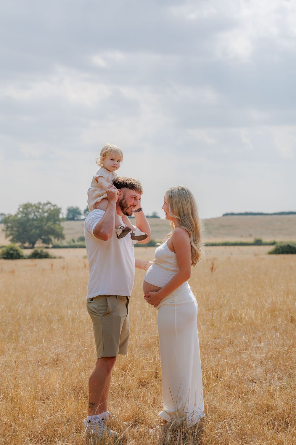 Pregnant woman in white dress with husband holding toddler in golden field under cloudy sky.