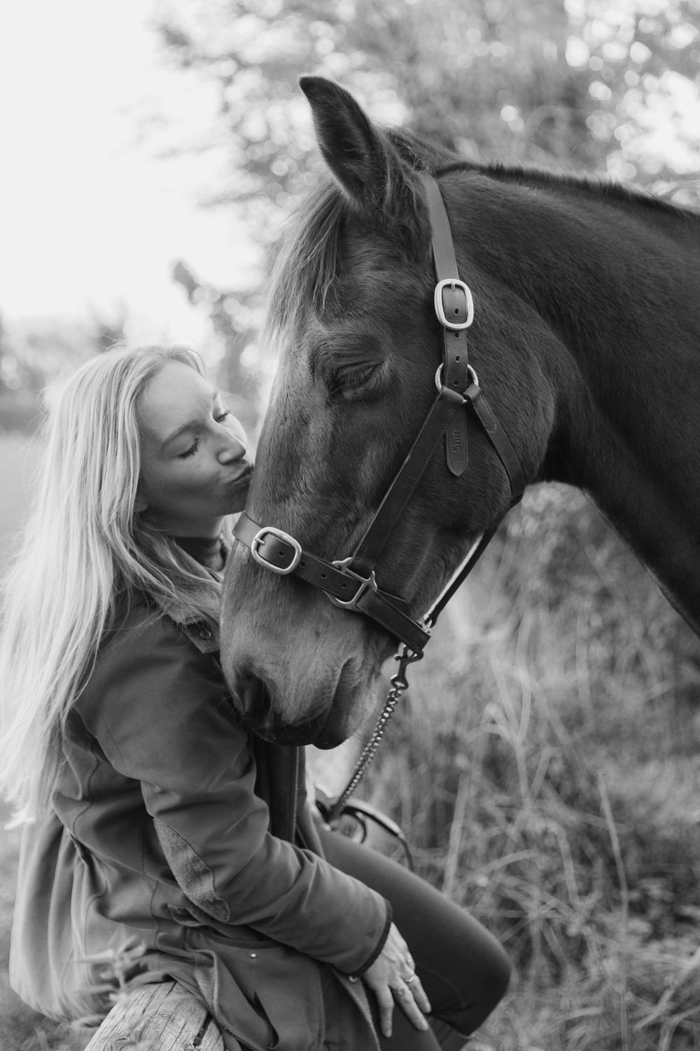 Black and white photo of a blonde woman kissing a horse wearing a bridle outdoors.