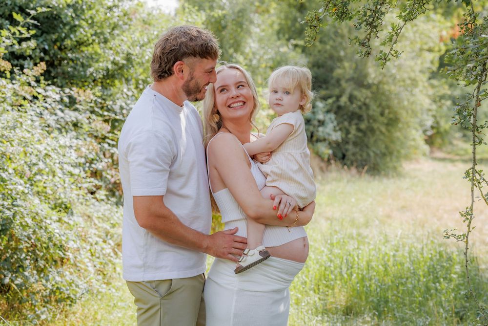 Happy pregnant couple with toddler in sunny green park setting, family maternity photo outdoors.