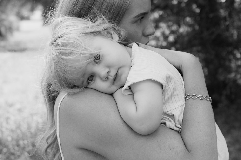 Black and white photo of a mother tenderly holding her sleepy young blonde toddler daughter close to her chest outdoors.