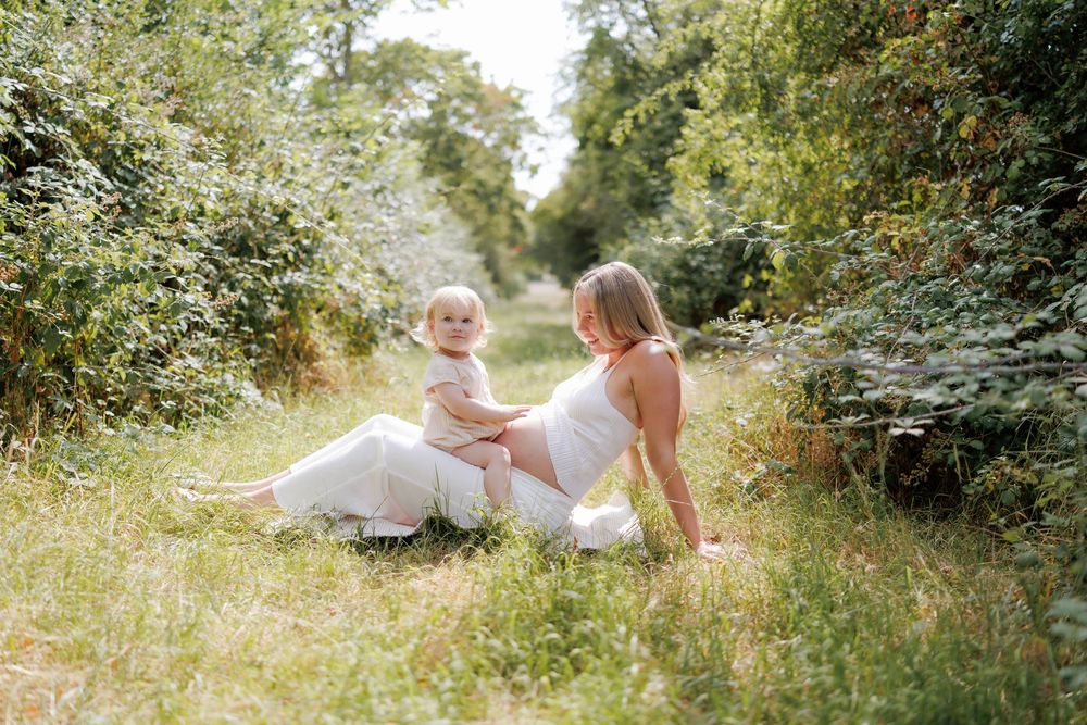 Pregnant woman in white dress sits outdoors in lush green meadow with a toddler on a sunny day.