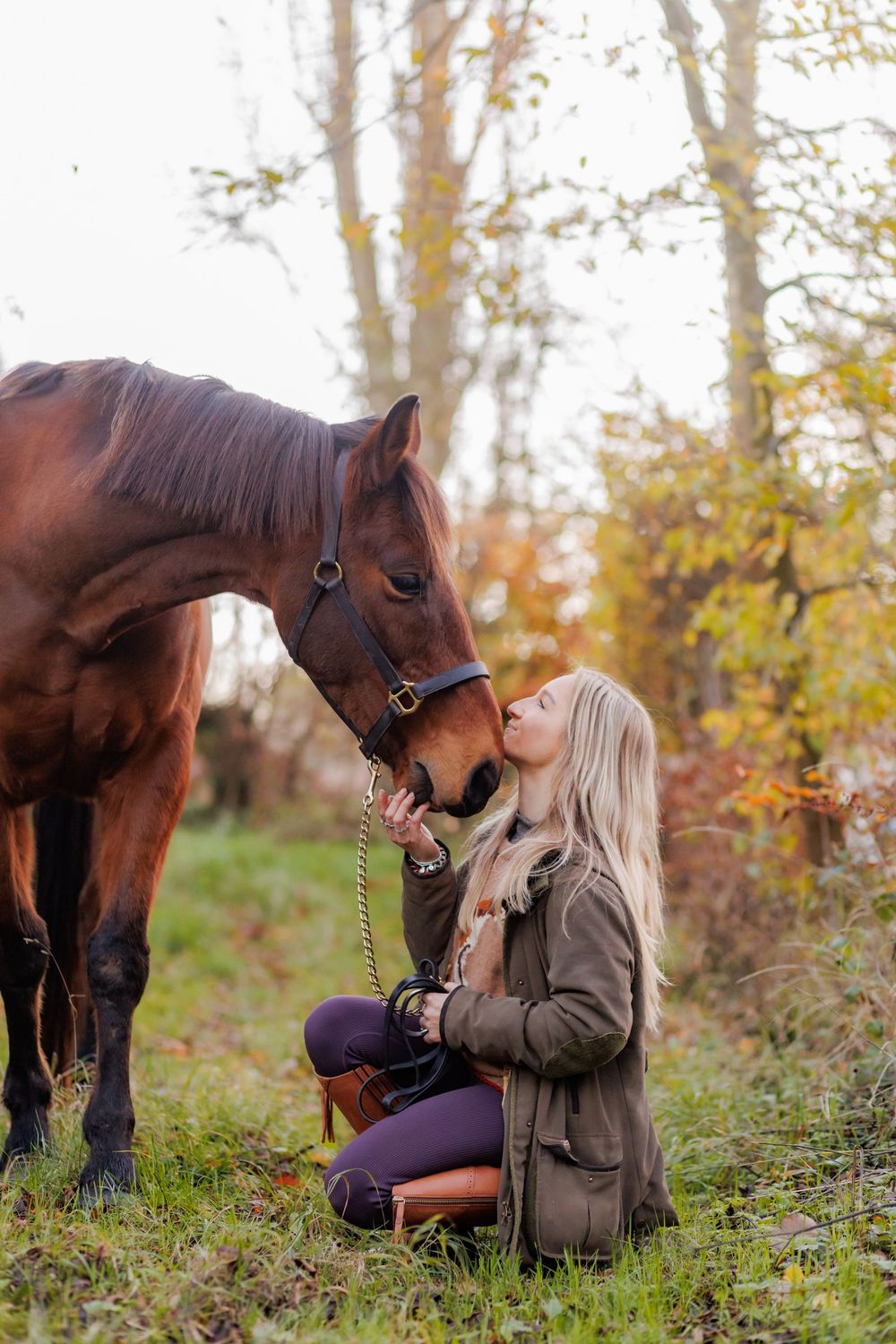 A blonde woman kneels in autumn forest, kissing a chestnut horse on the nose while holding its lead rope.