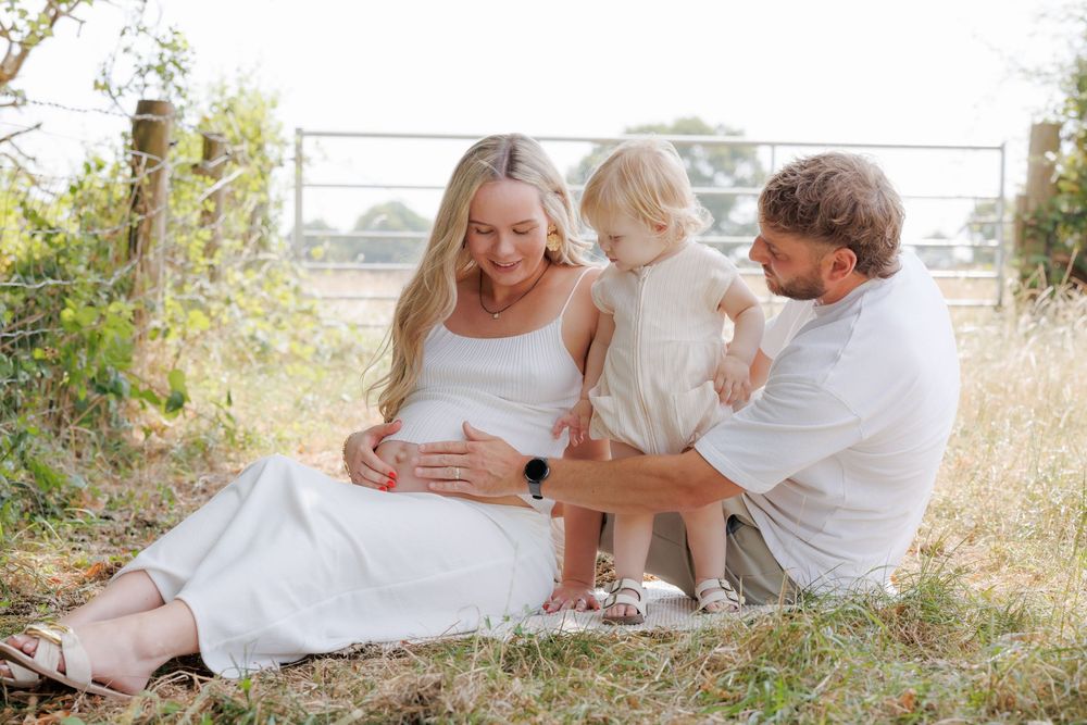 Pregnant woman in white dress sits outdoors with toddler and partner, enjoying a sunny family maternity photo session.