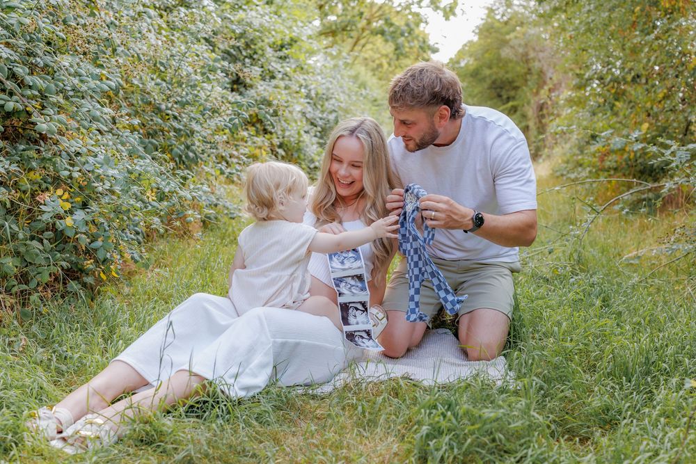 Pregnant couple sitting on grass with toddler reviewing ultrasound photos outdoors in lush green garden setting.