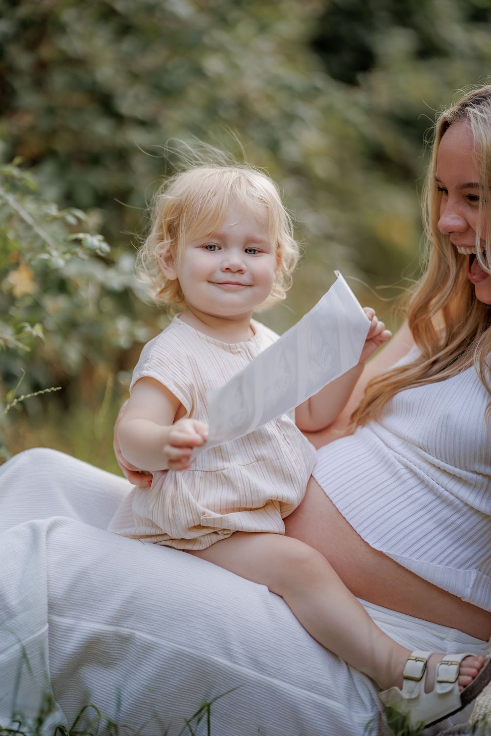 Toddler girl sitting on pregnant mother's lap outdoors, holding paper, smiling in natural garden setting.